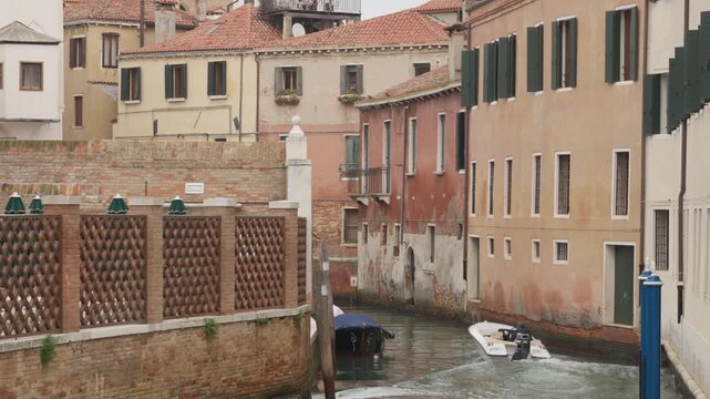 Small motorboat with outboard engine cruising through narrow Venetian canal. Traditional residential buildings with tile roofs and waterway in Venice Italy. Local urban transport and travel.