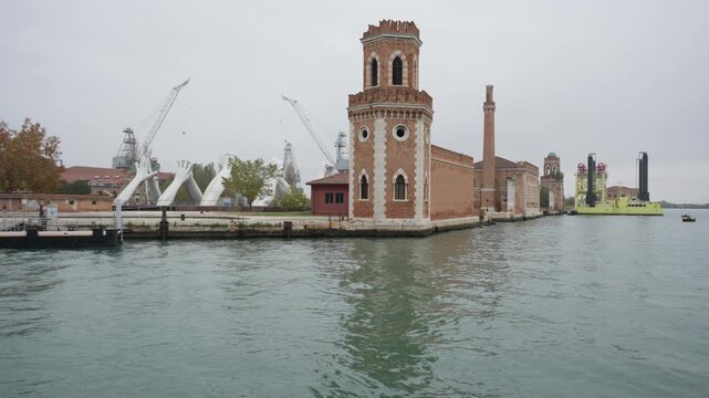 Giant white hand sculptures and historical Arsenale towers in Venice Italy. Scenic lagoon waterway view with brick architecture, industrial cranes and maritime vessels on a cloudy day.
