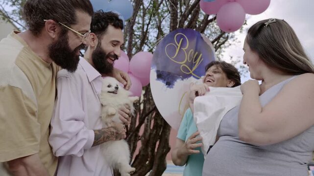 Happy family and friends celebrating a gender reveal party outdoors with pink and blue balloons, featuring a pregnant woman and a small white dog.