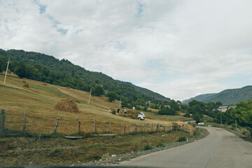 Rural road winds through rolling countryside with hay bales and wooden fences under a cloudy sky, capturing a peaceful agricultural landscape and expansive open space.