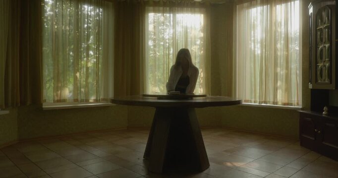 Girl enters sunlit spacious room and sits at round wooden table with single leg
