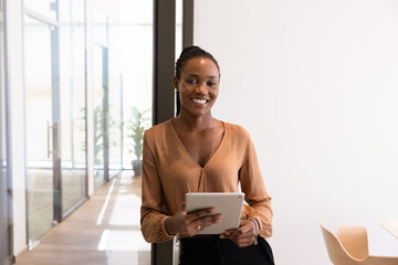 Positive confident young African American businesswoman standing in modern office workspace, holding digital tablet for managing online business process, looking at camera, posing for portrait