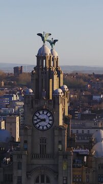 Aerial dawn view in Liverpool shows the Royal Liver Building clock, turrets, and Liver Birds, with warm light, long shadows, rooftops, and distant hills.