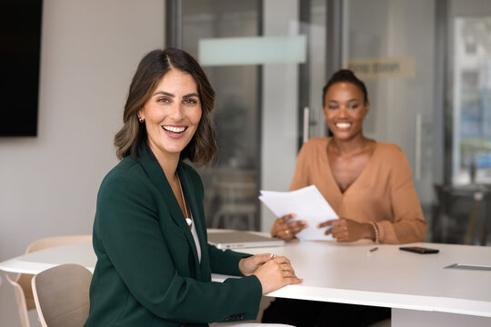 Happy confident Latin applicant woman sitting at meeting table with interviewer, recruit manager on job interview, looking at camera, smiling, posing for portrait, getting hired