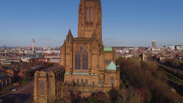 Aerial drone shot circles Liverpool Anglican Cathedral at sunrise, then pulls back to show Radio City Tower, Metropolitan Cathedral, winter trees, and waterfront.