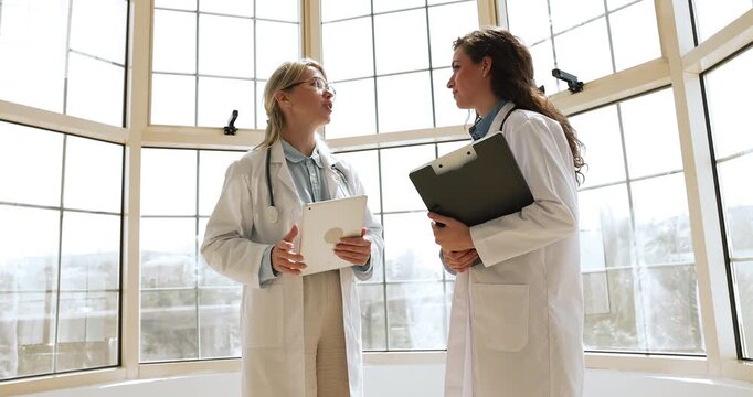 Two female doctors holding tablet and clipboard, shake hands standing near large windows in clinic, discuss patient case or diagnosis, exchanging opinions, concluding meeting with handshake gesture