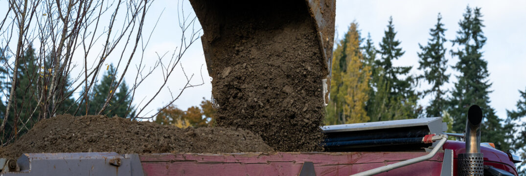 Excavator bucket attachment spilling dirt into a dump truck to haul away, public infrastructure construction site
