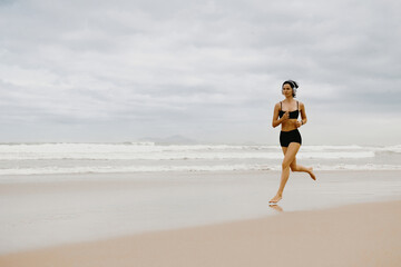 Young asian female running on beach in swimwear with cloudy sky and waves