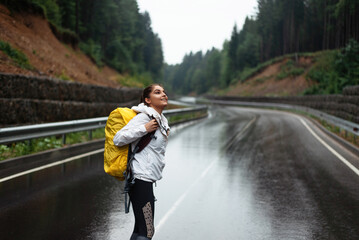 Young woman with a yellow backpack stands on a wet road looking up at the sky