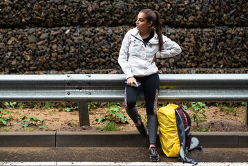 Young woman in athletic wear pauses by a gabion wall with backpack and phone