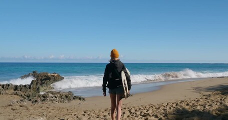 Woman walks on a sandy tropical beach next to ocean, holding a surfboard. 