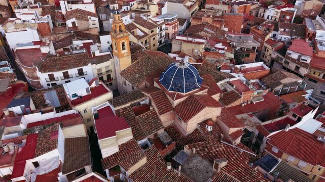 Aerial orbit around a historic church in Serra village as pigeons lift from tiled rooftops, revealing Mediterranean architecture and timeless charm in Valencia, Spain