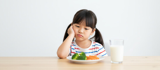 Unhappy little Asian girl refusing to eat healthy vegetables like broccoli and carrots with milk on table.