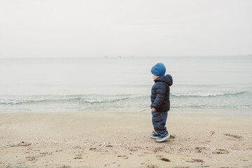 Child walks along sandy beach near water on a cloudy day in a coastal area with calm waves and no people around, focusing on the child's exploration of the surroundings
