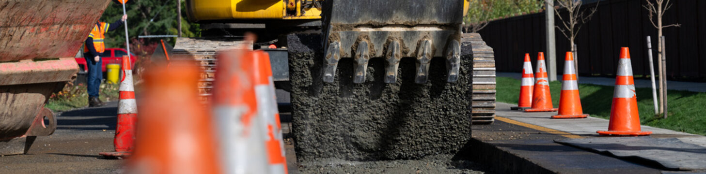 Excavator bucket attachment removing section of asphalt cut in road to dig a utilities trench, public infrastructure construction site, workman watching the project

