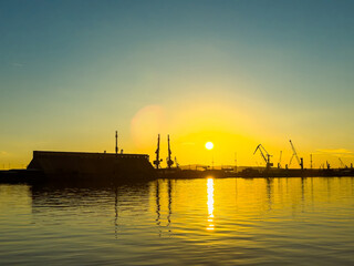 Fototapeta premium Sunset view over a harbor with cranes and a silhouette of a building along the water in the evening