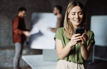 Young businesswoman using a smartphone in a modern office, with her professional team working in the blurred background