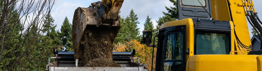Excavator bucket attachment spilling dirt into a dump truck to haul away, public infrastructure construction site
