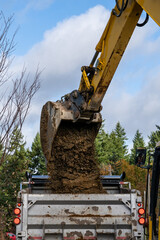 Excavator bucket attachment spilling dirt into a dump truck to haul away, public infrastructure construction site
