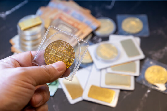 Hand holding real gold coin in protective case with scattered gold bars and banknotes background savings investment concept close-up