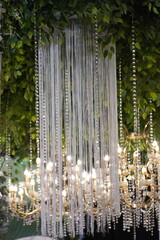 Elegant wedding decoration featuring a white fringe curtain and crystal beads hanging from a lush green foliage ceiling, with a glowing golden chandelier in a soft-focus background
