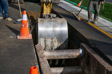 Excavator bucket attachment moving section of asphalt cut out of road to dig a utilities trench, workman using broom to sweep dirt back into hole, public infrastructure construction site
