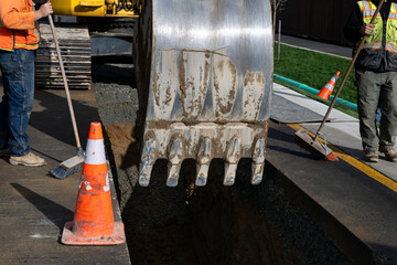 Excavator bucket attachment moving section of asphalt cut out of road to dig a utilities trench, workman using broom to sweep dirt back into hole, public infrastructure construction site
