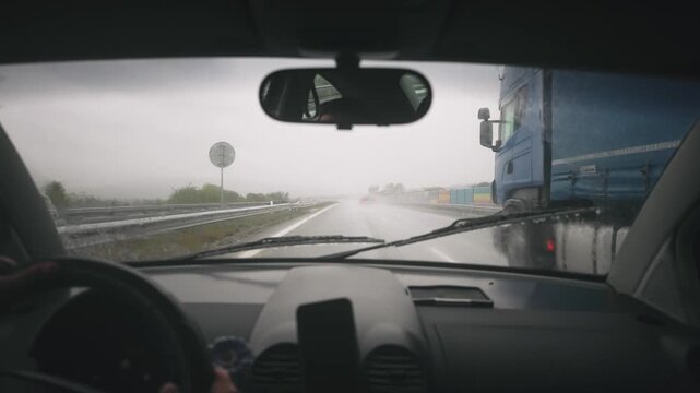 Pov of driving car overtaking an industrial truck with trailer transporting goods on rainy highway, first person view through windshield window, slow motion