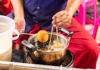 A street food vendor uses long wooden skewers to deep fry golden-brown dough balls in a pot of hot oil
