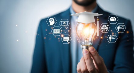Close up of a hand holding a glowing lightbulb with graduation cap and pixelated icons, symbolizing educational technology on gray background. Sparks around bulb, blue suit visible.