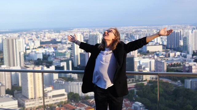 Joyful businesswoman in a suit jumping with arms outstretched on a skyscraper rooftop, celebrating success and freedom with a beautiful city skyline view during a sunny, windy day