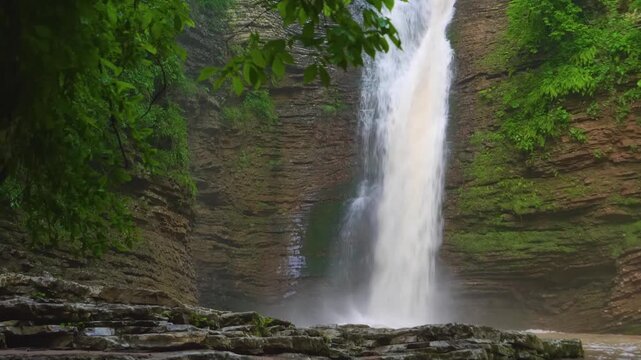 A picturesque view of one of the Rufabgo waterfalls located in the Republic of Adygea, Russia. The powerful cascade flows over layered rock formations amidst a vibrant, green forest landscape.