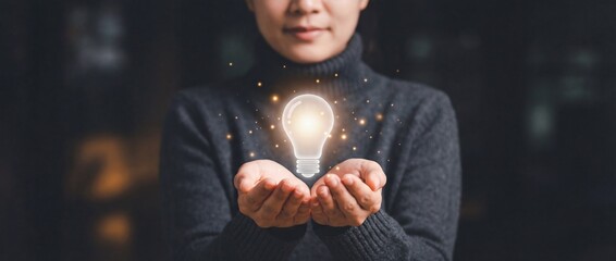 Woman in dark sweater holding glowing virtual light bulb with golden sparkles representing new idea