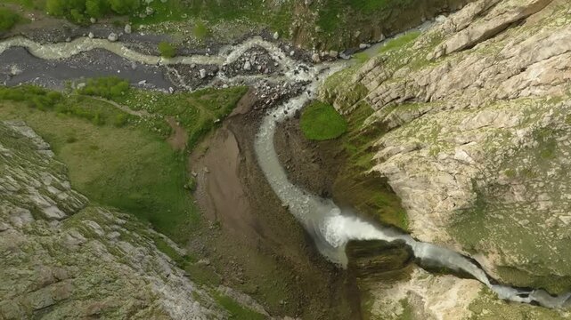 Drone footage captures a breathtaking, high-angle view of a vibrant, winding river and a small waterfall cascading through the dramatic, rugged landscape of the Kara-Syrt Ridge in the Republic of Dage