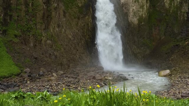 A powerful stream of water cascades down a rugged, rocky cliff face in the scenic Kara-Syrt Ridge area of Dagestan, within the beautiful Caucasus Mountains. Lush green vegetation and small yellow flow