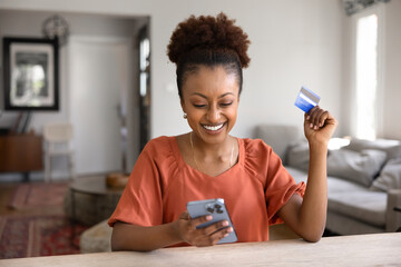 Cheerful excited African American bank client woman using credit card for online shopping, getting...