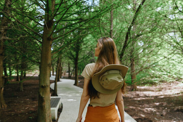 Woman walking on forest path with hat attached to backpack, long hair and casual clothes, enjoying green trees and shaded trail in nature park during summer outdoor stroll.