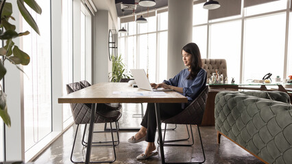 Positive young Asian manager woman working at desktop in office co-working space alone, typing on laptop, analyzing Internet sales for marketing strategy. Side view wide banner shot