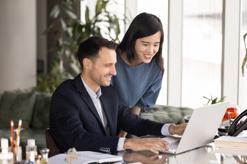 Two positive multiethnic business colleagues discussing successful start of company product, Internet sales growth, talking at office workplace, looking at laptop display, speaking, smiling