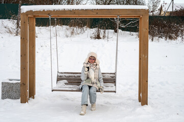 Woman in winter clothes with a cup of coffee on a swing in a snowy park.