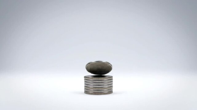 A stone balances delicately atop a stack of coins on a plain white surface from a straightforward viewpoint