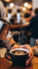 Close-up of a person's hand holding a black coffee cup as steam rises while pouring hot water from a metal kettle in a blurred cafe background with warm tones.