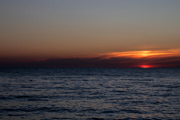 Obraz premium The reflection of the setting sun on the waves. A view of the sea at sunset. The sky over the sea at sunset. In the distance, bright sunbeams and reflections of the warm light of the setting sun glist