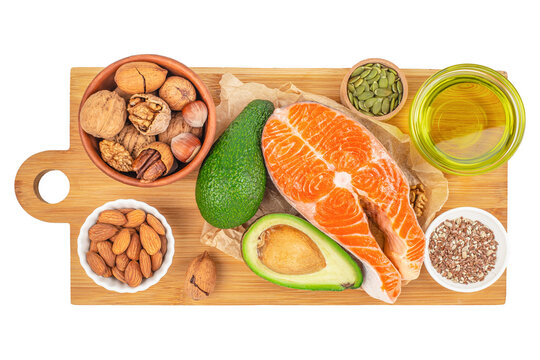 Top view of healthy fats food sources including raw salmon steak, avocado, nuts, seeds and olive oil arranged on wooden cutting board, balanced nutrition and clean eating concept