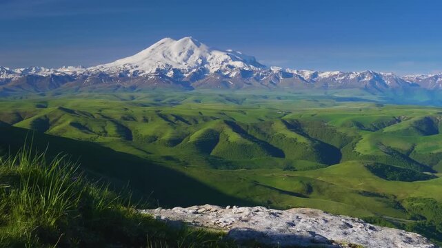 panoramic landscape of the snow-capped peaks of Mount Elbrus, the highest mountain in Europe, viewed from the vast green rolling hills of the Bolshoy Bermamyt plateau in the early morning.