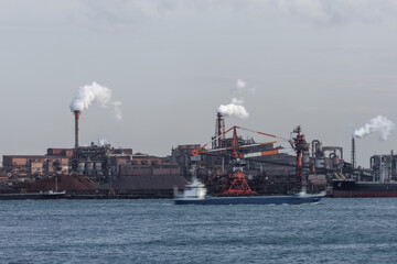 Fototapeta premium Motion blurred cargo ship passes by an industrial waterfront complex backdrop featuring docked ships, raw materials, multiple smoke emitting chimneys, and cranes and various freight equipment.