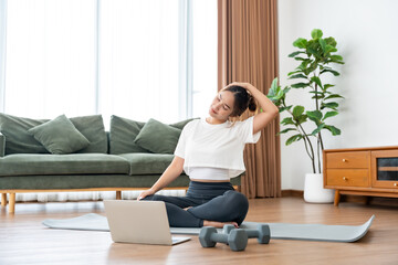 Young Asian woman stretching yoga workout on exercise mat while online training class with computer laptop in living room