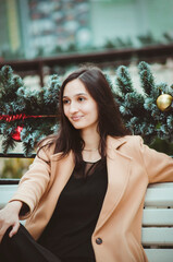 A beautiful young woman with long dark hair, wearing a black blouse, trousers, and a beige coat, sits on a park bench. Vertical photo.