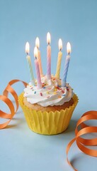 birthday cupcake with lit candles and sprinkles on blue background with orange ribbon streamers, festive party dessert, celebration concept with copy space