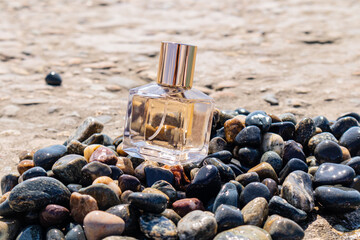 Glass perfume bottle with gold cap on a pebble beach with the sea in the background. Presentation of the summer fragrance of luxury perfume.